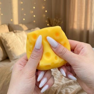 May include: A yellow cheese-shaped stress ball with several holes. The stress ball is held between two hands with long, white fingernails. The background is blurred, with soft lighting and neutral tones.
