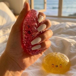May include: A translucent pink squishy toy filled with glitter and a printed smiling face, held in a palm, alongside a second translucent yellow squishy toy with a similar smiling face, both featuring rounded shapes and a jelly-like texture.