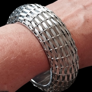 May include: A wide, silver-toned cuff bracelet featuring a detailed woven wire pattern that mimics the appearance of a basket weave. The metallic surface has a polished finish, creating a reflective texture across the entire exterior of the band.