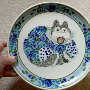May include: A white ceramic plate with a blue and green floral border. The center of the plate features a hand-painted illustration of a gray cat wearing a blue and white floral dress and holding a pink flower.