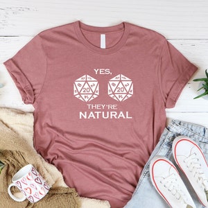 Puede incluir: Una camiseta color malva con un gráfico blanco de dos dados de 20 caras y el texto "YES, THEY'RE NATURAL".