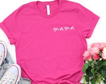 以下が含まれることがあります： ピンクのTシャツに、白で「mama」と書いてあります。Tシャツは白い背景に置かれ、白いスニーカーとピンクの花束が添えられています。