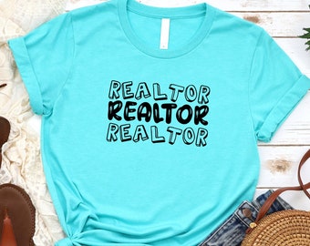 May include: A turquoise blue t-shirt with the text "REALTOR" repeated three times in black. The shirt is laid out on a white wooden surface with a pair of brown sandals, a white crocheted shawl, a straw bag, and a green succulent plant.