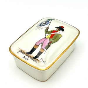 May include: A white porcelain box with a gold trim and a painted image of a soldier in a green and red uniform holding a flag. The box has the text "Garde Imperiale 1811" written on the bottom.