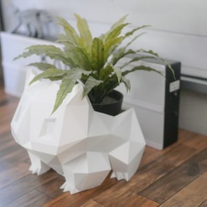 Könnte beinhalten: Eine weiße, geometrische Skulptur im Origami-Stil eines Comic-Tieres, möglicherweise eines Hundes oder eines Fuchses, mit einer grünen, belaubten Pflanze in einem schwarzen Topf davor.