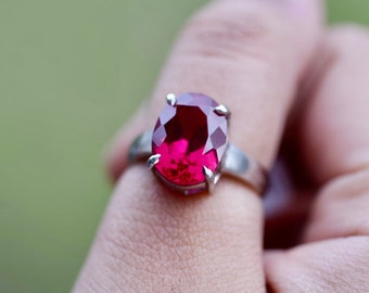May include: A silver ring with a large, oval-shaped, deep red gemstone.