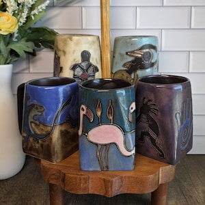 Könnte beinhalten: Ein Set aus fünf Keramikbechern mit verschiedenen handbemalten Motiven wie einem rosa Flamingo, einer Schildkröte, einem Flötenspieler und Tierfiguren, angeordnet auf einem hölzernen Etagenständer mit einem zentralen Griff.