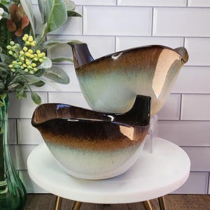 May include: Two ceramic serving bowls with a unique design. The bowls have a brown, green, and white gradient color scheme. Each bowl has a handle. The bowls are on a small white table with gold legs. A green vase with flowers is in the background.