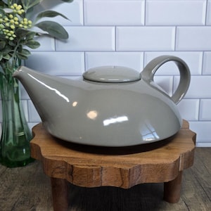 May include: A gray ceramic teapot with a matching lid and a curved handle. The teapot sits on a small, round, wooden pedestal with a scalloped edge. A green glass vase with flowers is in the background.