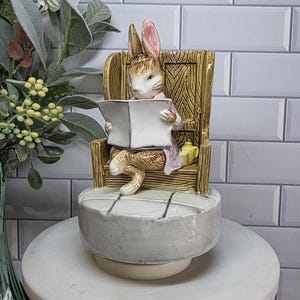 Peut inclure: Figurine en céramique représentant un lapin lisant un livre, assis dans une chaise en bois. Le lapin porte une veste rose et est posé sur une base ronde grise. Un vase vert avec des fleurs blanches et de la verdure est en arrière-plan.