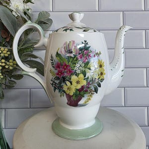 Pode incluir: Bule de cerâmica branco com um design de buquê floral. O bule apresenta uma alça e base verdes e uma tampa com um botão em forma de flor. O buquê inclui flores rosa, amarelas e vermelhas.