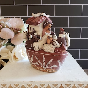 Könnte beinhalten: Eine Keramikfigur, die Noahs Arche mit einem braunen und weißen Design darstellt. Die Arche ist mit Tieren gefüllt, darunter ein Hahn, ein Schaf, eine Kuh, ein Esel und ein Schwein. Die Arche hat ein kleines Haus oben mit einem Dach und zwei Vögeln auf dem Dach.