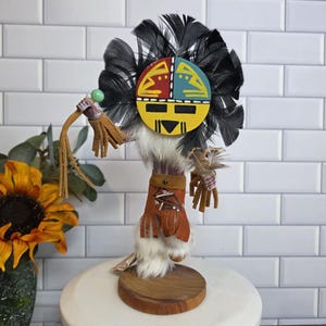 May include: A Native American-style doll with a painted face in yellow, red, and blue, adorned with black feathers. The doll has a fur body, leather clothing, and is holding a green sphere and a fringed whip. It stands on a wooden base.