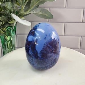 Op de afbeelding: Een blauw en paars eivormig kristal met een draaiend patroon. Het kristal ligt op een wit oppervlak.