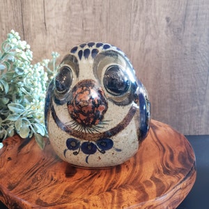 May include: A ceramic bird figurine with blue, brown, and white painted details. The bird has large eyes and a beak with a copper-colored glaze. The figurine is sitting on a wooden base.