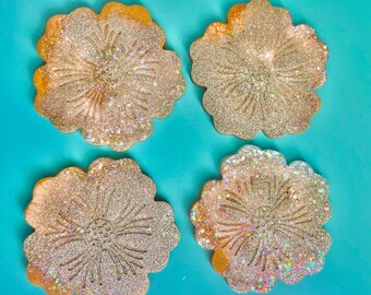 May include: Four flower-shaped coasters with a glittery, textured surface. The coasters have a light gold color with a floral design etched into the center. The background is a bright teal color.