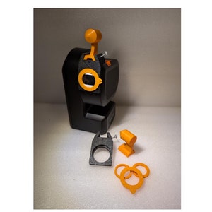 Puede incluir: Una impresora 3D negra y naranja con una pequeña boquilla naranja y una base negra y gris. La impresora está sobre una superficie blanca con un anillo negro y gris, un anillo negro y gris con un pequeño anillo naranja en el interior y un pequeño soporte naranja.