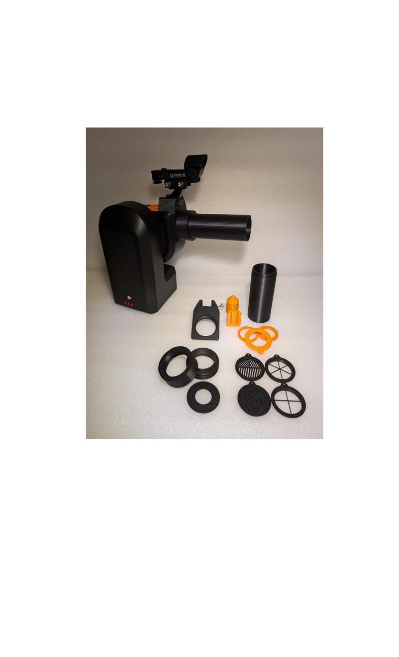 May include: A black projector with a lens and a small orange and black camera attached. The projector is surrounded by various black and orange accessories, including a lens cap, a filter, and a small orange piece.