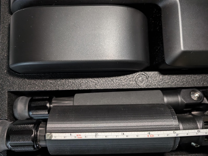 May include: A close-up of a black tripod with a ruler measuring 10 inches. The tripod is made of metal and has a black rubber grip.