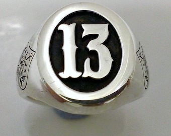 Può includere: Un anello d'argento con un designo circolare nero che presenta il numero "13" in bianco. L'anello ha un designo dettagliato sui lati.