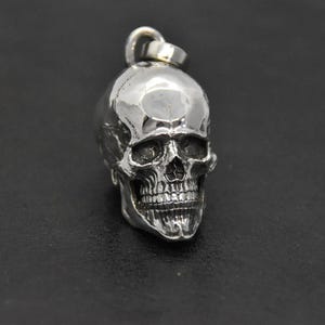 May include: A silver skull pendant with a detailed design. The skull is facing forward with a slightly open mouth.