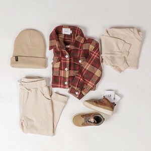 K&ouml;nnte beinhalten: Eine Flatlay eines Herbstoutfits f&uuml;r ein Kind. Das Outfit besteht aus einem rot-braunen karierten Flanellhemd, beigen Jogginghosen, einer beigen M&uuml;tze, einem beigen Langarmshirt, braunen High-Top-Sneakern und wei&szlig;en Socken.