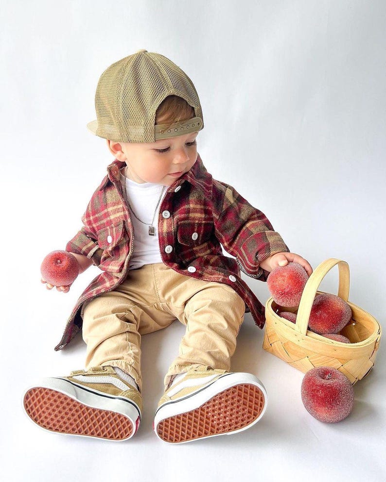 K&ouml;nnte beinhalten: Ein kleines Kind tr&auml;gt ein rot-schwarzes Karohemd, eine braune Netzbaseballkappe und eine beige Hose und sitzt auf einer wei&szlig;en Oberfl&auml;che. Das Kind h&auml;lt einen roten Pfirsich in der einen Hand und einen Korb mit Pfirsichen in der anderen. Das Kind tr&auml;gt beige Turnschuhe.