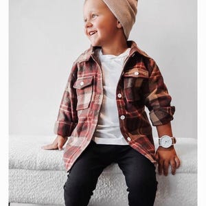 K&ouml;nnte beinhalten: Ein kleiner Junge tr&auml;gt ein rot-beiges kariertes Flanellhemd, eine schwarze Hose mit wei&szlig;en Streifen, ein wei&szlig;es T-Shirt und eine beige M&uuml;tze. Er sitzt auf einer wei&szlig;en Bank und l&auml;chelt.