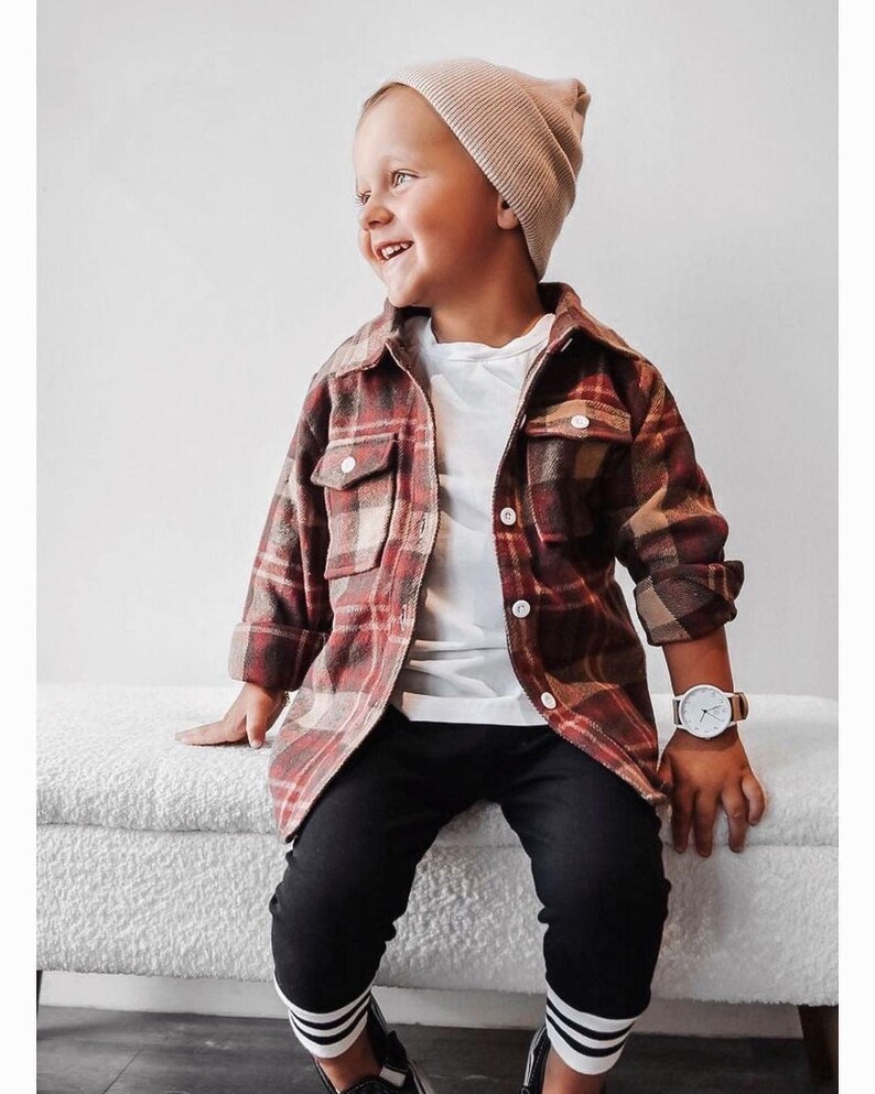 K&ouml;nnte beinhalten: Ein kleiner Junge tr&auml;gt ein rot-beiges kariertes Flanellhemd, eine schwarze Hose mit wei&szlig;en Streifen, ein wei&szlig;es T-Shirt und eine beige M&uuml;tze. Er sitzt auf einer wei&szlig;en Bank und l&auml;chelt.