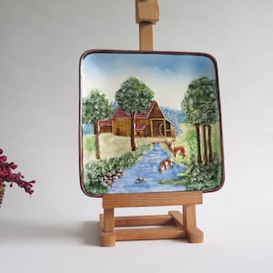 May include: Square ceramic decorative plate depicting a rural landscape. The plate shows a house, trees, a river, and deer. The plate is displayed on a small wooden easel.