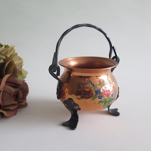 Puede incluir: Una pequeña olla de cobre martillado con un asa y tres patas de metal negro. La olla presenta un diseño floral pintado a mano en rojo, azul y verde, con la palabra Rättvik escrita en letra cursiva negra sobre la superficie de cobre.