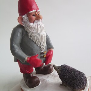 May include: A red-hatted gnome with a gray coat and red mittens looks at a small gray hedgehog. The gnome is standing on a white base with the text "NWF 2007" on it.