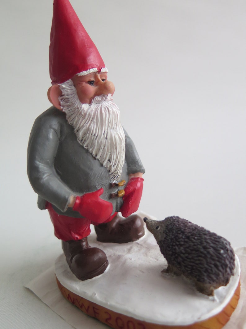 May include: A red-hatted gnome with a gray coat and red mittens looks at a small gray hedgehog. The gnome is standing on a white base with the text "NWF 2007" on it.
