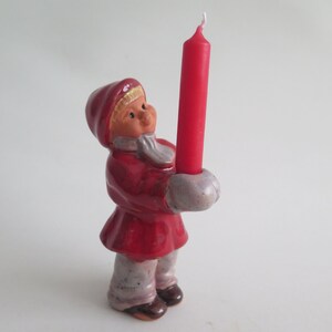 May include: A ceramic figurine of a child in a red coat and hood, holding a tall red candle. The figurine has a light skin tone, a white scarf, and gray mittens. The candle is approximately 6 inches tall.