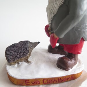 May include: A gray and red gnome figurine with a white beard stands on a wooden base with the text "Eva Dahlberg". A small brown hedgehog figurine is in front of the gnome.