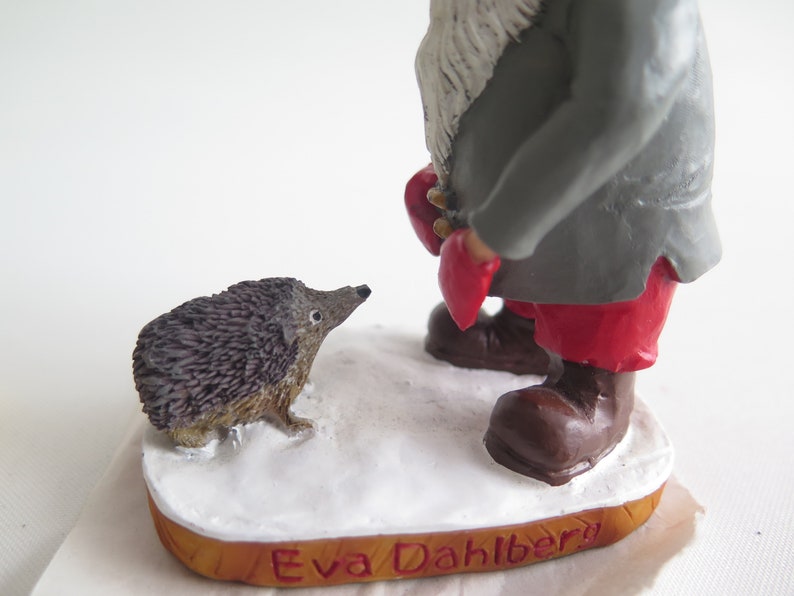 May include: A gray and red gnome figurine with a white beard stands on a wooden base with the text "Eva Dahlberg". A small brown hedgehog figurine is in front of the gnome.