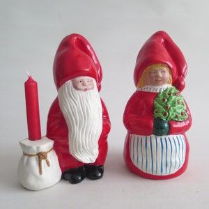 Puede incluir: Dos figuras navideñas de cerámica. Una es una figura de Papá Noel con una larga barba blanca, traje rojo y un portavelas. La otra es una figura femenina que sostiene un árbol de Navidad. Ambas figuras llevan sombreros y trajes rojos.