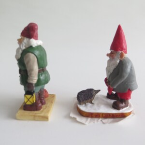May include: Two small gnome figurines, one in green and red with a lantern, the other in gray and red with a hedgehog. Both are standing on wooden bases.