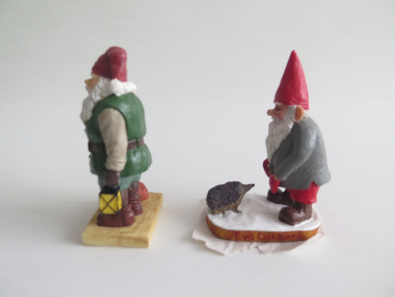 May include: Two small gnome figurines, one in green and red with a lantern, the other in gray and red with a hedgehog. Both are standing on wooden bases.