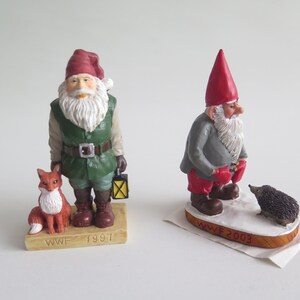 May include: Two small, hand-painted figurines of gnomes. The gnome on the left is wearing a green jacket, red hat, and holding a lantern. He is standing next to a small, orange fox. The gnome on the right is wearing a gray jacket, red hat, and red mittens. He is standing next to a small, brown hedgehog. Both gnomes are standing on a wooden base with the text "WWF" and the year. The gnome on the left is from 1997 and the gnome on the right is from 2003.