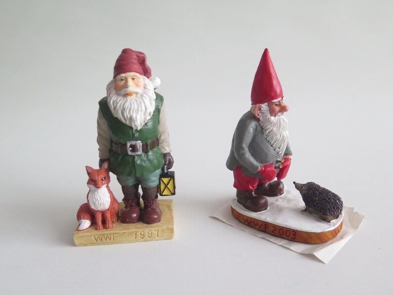 May include: Two small, hand-painted figurines of gnomes. The gnome on the left is wearing a green jacket, red hat, and holding a lantern. He is standing next to a small, orange fox. The gnome on the right is wearing a gray jacket, red hat, and red mittens. He is standing next to a small, brown hedgehog. Both gnomes are standing on a wooden base with the text "WWF" and the year. The gnome on the left is from 1997 and the gnome on the right is from 2003.