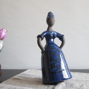 May include: A blue ceramic figurine of a woman wearing a blue dress and a head covering. The figurine is standing on a white surface.
