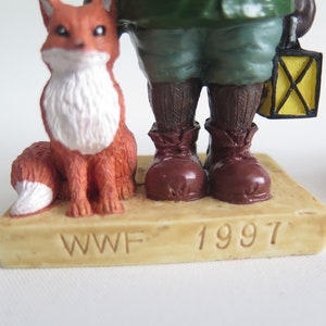 May include: A brown figurine of a person wearing a green jacket and brown boots, holding a yellow lantern. A red fox figurine sits beside the person. The base of the figurine is beige and reads "WWF 1997".
