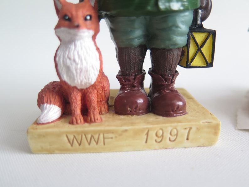 May include: A brown figurine of a person wearing a green jacket and brown boots, holding a yellow lantern. A red fox figurine sits beside the person. The base of the figurine is beige and reads "WWF 1997".