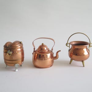 Puede incluir: Tres miniaturas de utensilios de cocina de cobre: un recipiente en forma de barril, una tetera y un pequeño caldero con asa. Los artículos se exhiben sobre un fondo blanco liso, mostrando sus superficies brillantes y reflectantes.