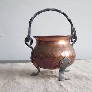 Puede incluir: Un pequeño caldero de cobre martillado con un asa de hierro forjado negro. El caldero se asienta sobre tres patas de hierro forjado.