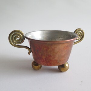 May include: A small, antique copper bowl with brass accents. The bowl has a hammered texture and a silver interior. It features two ornate, swirled handles and rests on three small, round feet. The copper has a reddish-brown patina.