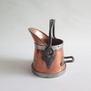 May include: A small copper pitcher with a handle and spout. The pitcher has a copper body with a silver-colored metal band around the top and bottom. A dark metal handle is attached to the top, and a small handle is on the side.