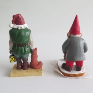 May include: Two Christmas gnome figurines. One gnome is wearing a green jacket, red hat, and holding a lantern and a fox. The other gnome is wearing a gray jacket and red hat. Both gnomes are standing on a wooden base.