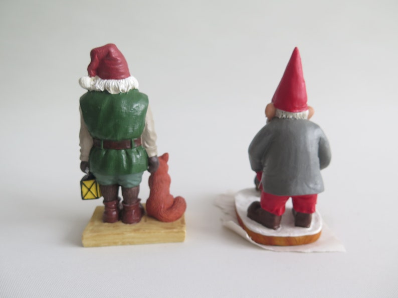 May include: Two Christmas gnome figurines. One gnome is wearing a green jacket, red hat, and holding a lantern and a fox. The other gnome is wearing a gray jacket and red hat. Both gnomes are standing on a wooden base.